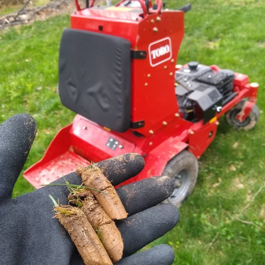 Gloved hand holding three freshly pulled soil cores in front of a red Toro stand-on aerator
