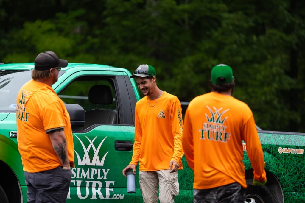 Three Simply Turf technicians in uniform laughing together next to a grass-wrapped Simply Turf truck